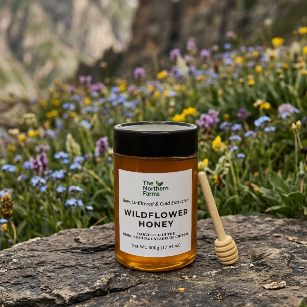 A jar of The Northern Farms Wildflower Honey (Jangli shehad) sits on a dark stone surface with a wooden honey dipper beside it, set against a blurred mountain meadow background filled with purple, blue, and yellow wildflowers.