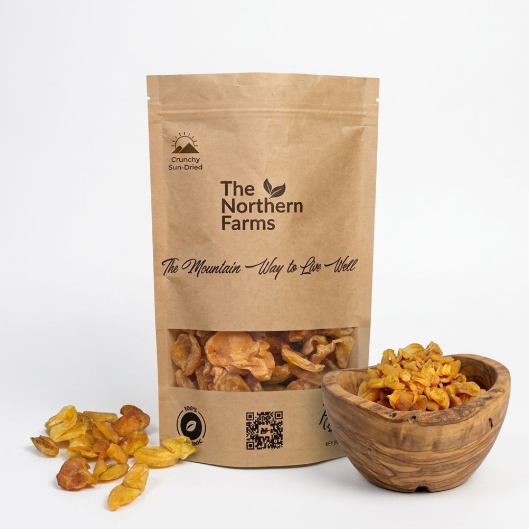 A stand-up kraft paper pouch of dried Apricots (Khubani) from The Northern Farms, shown on a white background with loose dried khurmani pieces beside it and a small wooden bowl filled with the halves of sun dried organic apricots from Chitral Valley
