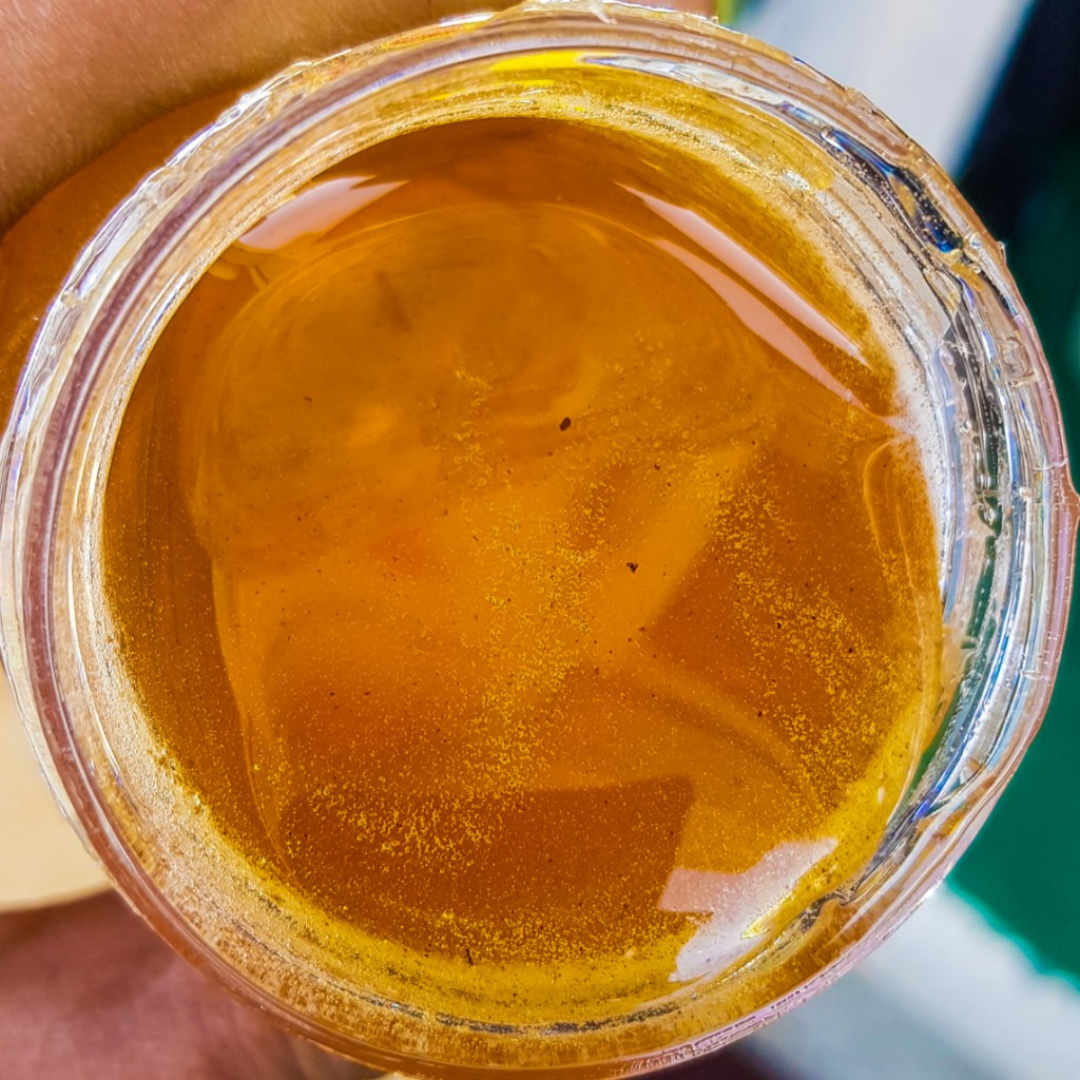 A close-up top-down view of a clear jar filled with The Northern Farms' Russian Olive Forest honey, held by a hand. The honey has a warm, translucent yellow-orange color with visible fine particles and light reflections.